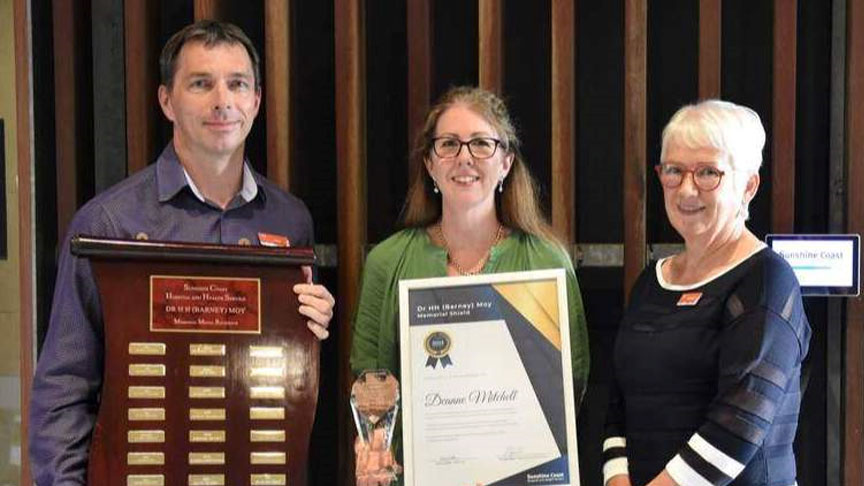 Three people stand together holding a trophy, plaque and certificate