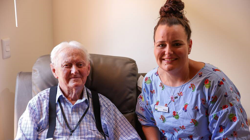 A 94-year-old man sat in a chair with a nurse crouching down next to him.