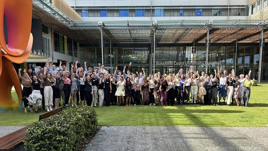 A large group of people standing on the grass outside Sunshine Coast University Hospital with arms raised in celebration.