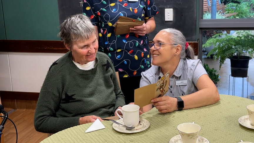 Two people sat at a table with one showing the other a Christmas card. A third person stands behind them with a handful of envelopes.