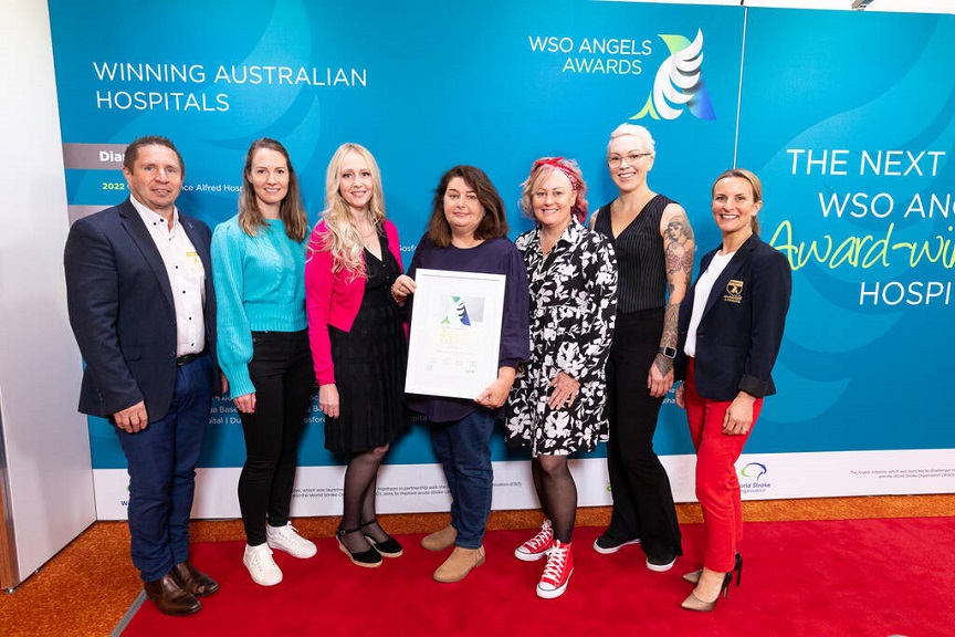 Seven people standing on a red carpet in front of a blue WSO Angels Awards banner holding a certificate