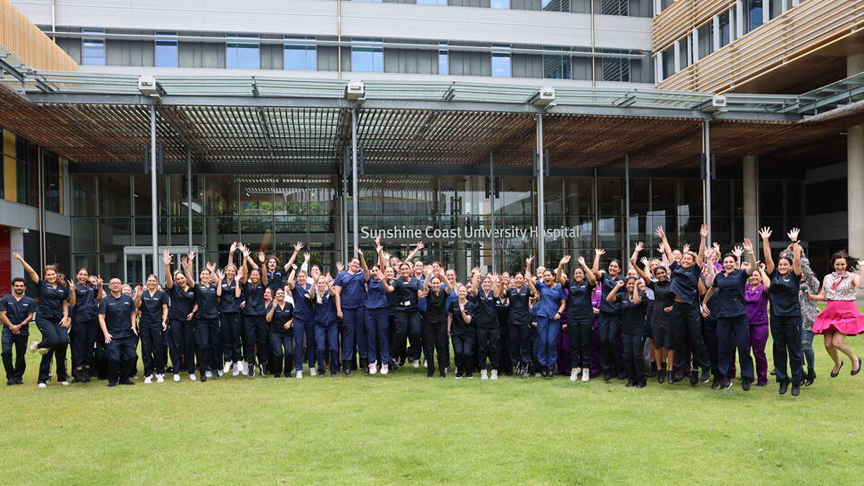 A large group of people jumping and raising their hands in the air on the grass outside Sunshine Coast University Hospital