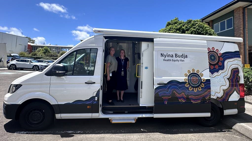 A white van with Aboriginal and Torres Strait Islander artwork on the side, parked in a car park with the door open and 2 people standing inside.