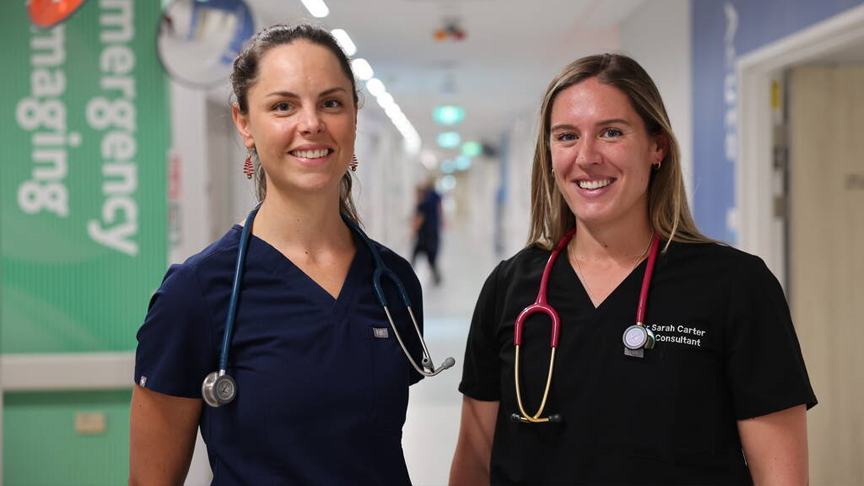 A doctor and nurse standing in the hallway of an emergency department wearing scrubs and stethoscopes.