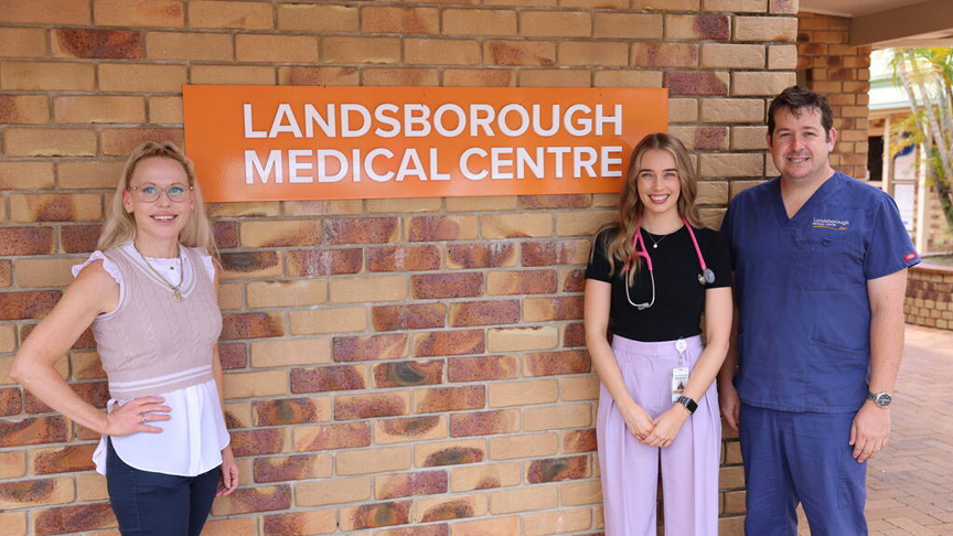 Three people stand in front of a brick wall. One to the left and two to the right of an orange sign on the wall with white writing that says Landsborough Medical Centre.