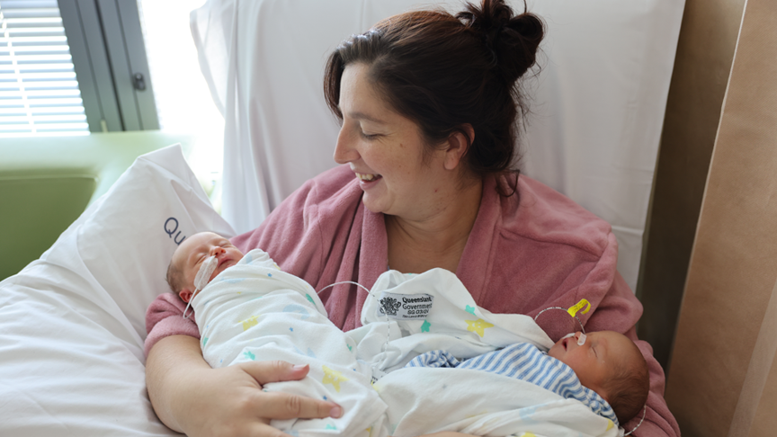 Mother sat on a chair holds her new twins, cradling one in each arm with her right arm supported by a pillow.