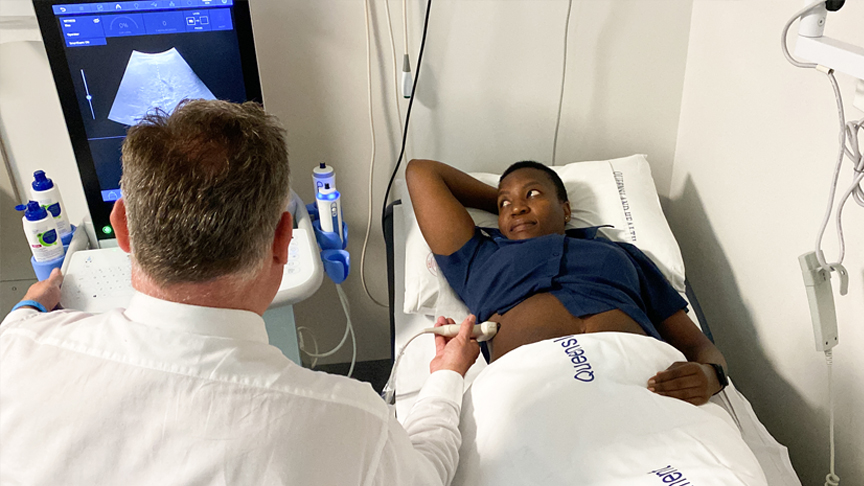 Professor James O’Beirne reviews an ultrasound image standing next to a patient lying on a bed 