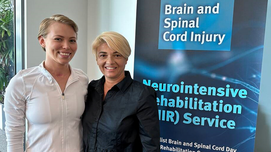 Two people stand in front of a blue banner for the Neurointensive Rehabilitation Service 